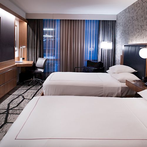 A modern hotel room with two neatly made beds, a wall-mounted TV, a desk, chair, and large windows with curtains, stylish decor and ample lighting.