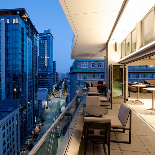A modern rooftop terrace with seating overlooks a cityscape of tall buildings at dusk, featuring sleek architectural lines and well-lit interiors.