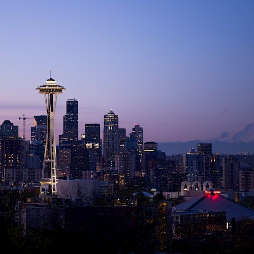 A twilight view of Seattle's skyline, featuring the Space Needle with Mount Rainier visible in the background, under a clear evening sky.