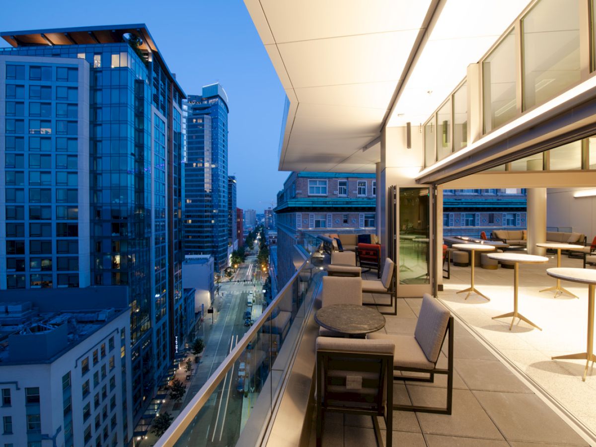 A modern balcony with seating and tables overlooks a city street lined with tall buildings during the evening.