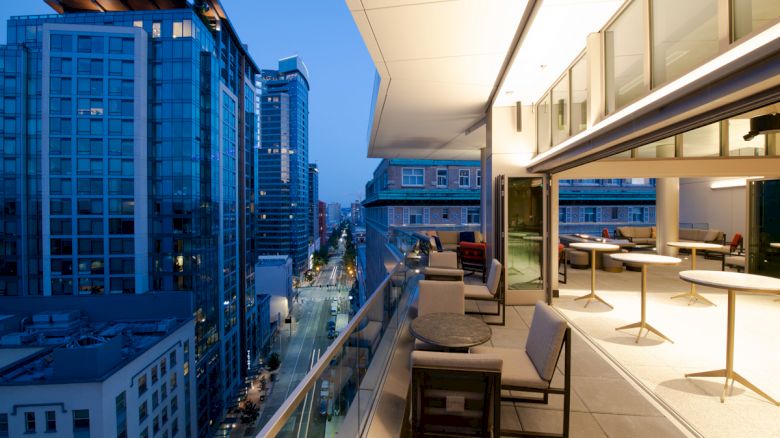 A modern balcony with seating and tables overlooks a city street lined with tall buildings during the evening.