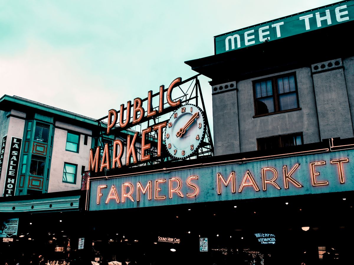 This image depicts a section of the exterior of a Public Farmers Market, showing neon signs and buildings under a cloudy sky with urban architecture.