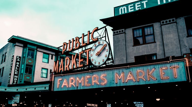 This image depicts a section of the exterior of a Public Farmers Market, showing neon signs and buildings under a cloudy sky with urban architecture.