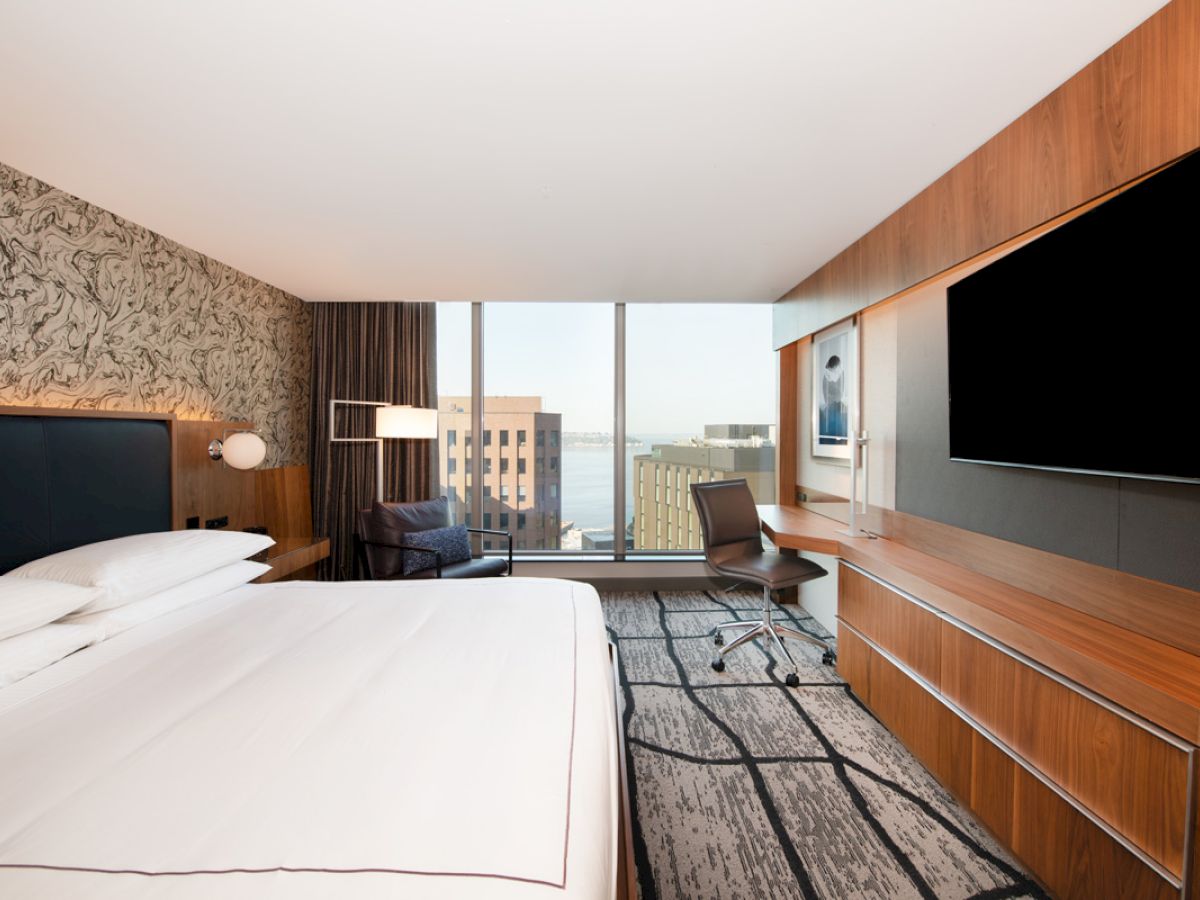 A modern hotel room with a king-sized bed, large TV, desk, and floor-to-ceiling windows offering a view of city buildings and water in the distance.