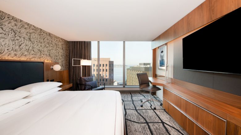A modern hotel room with a king-sized bed, large TV, desk, and floor-to-ceiling windows offering a view of city buildings and water in the distance.