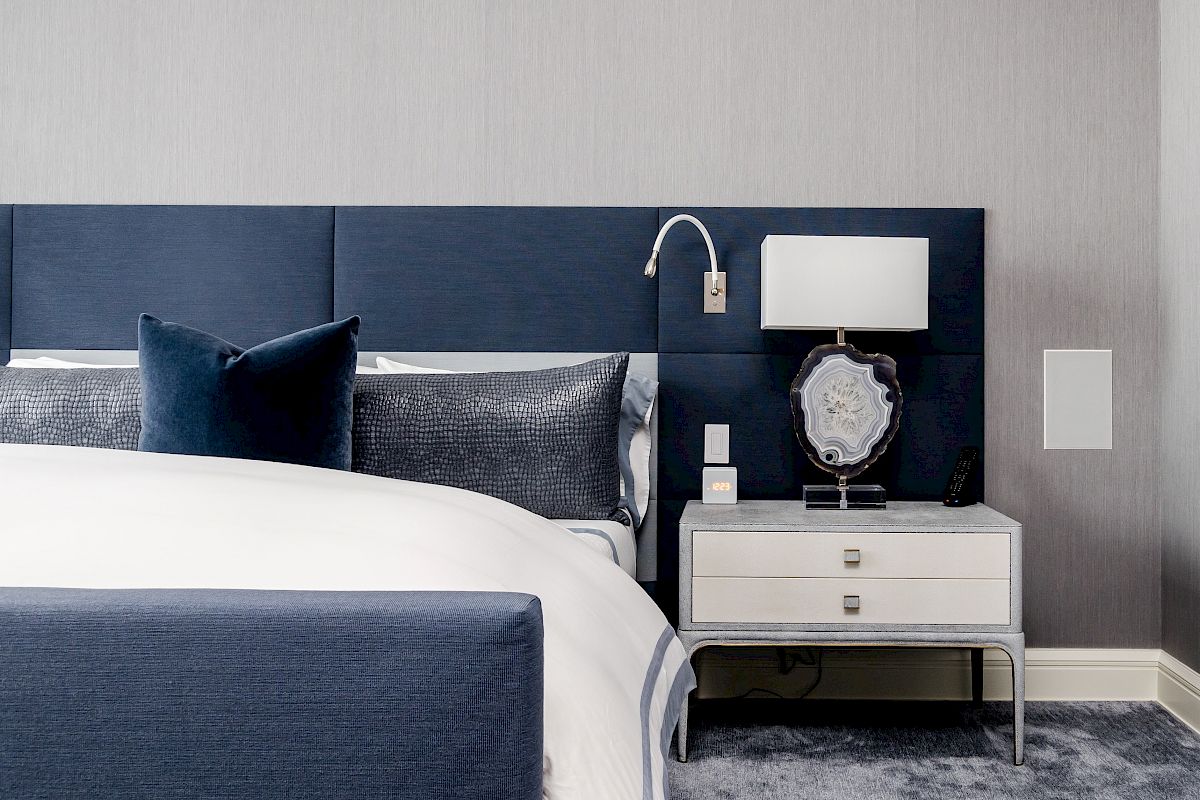 A modern bedroom with a blue upholstered bed, white bedding, and a nightstand with a lamp and decorative object, on a blue carpet.