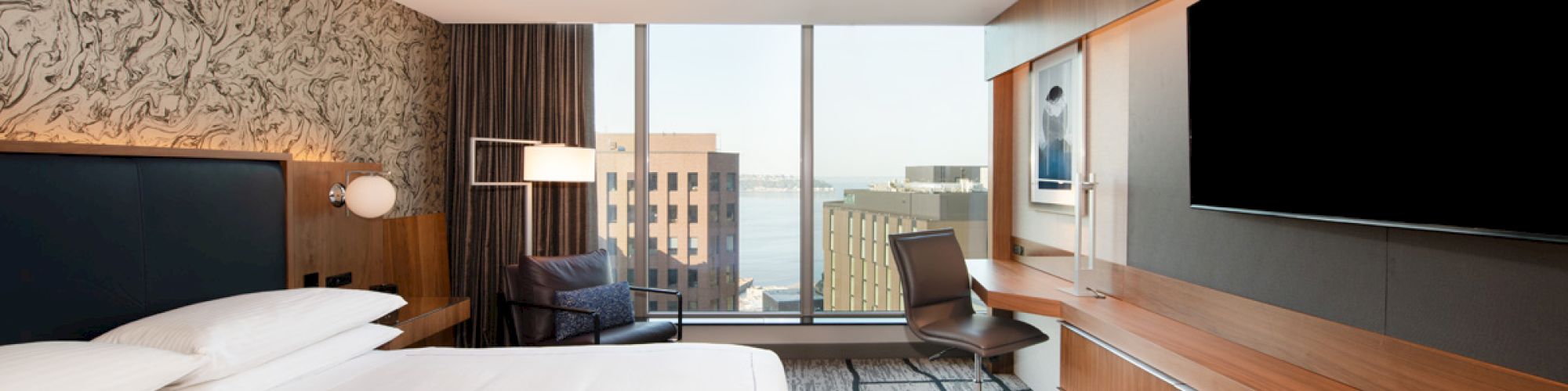 The image shows a modern hotel room with a large bed, wall-mounted TV, desk, chair, floor-to-ceiling window with a city view, and contemporary decor.